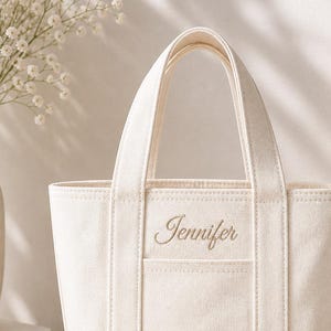 May include: Cream-colored canvas tote bag with a rectangular shape and two handles. The name "Jennifer" is embroidered in a cursive font on the front pocket. The bag is sitting on a wooden surface.