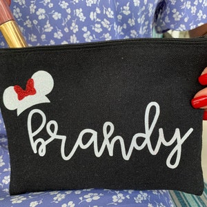 May include: Black canvas cosmetic bag featuring a white Minnie Mouse design and the name "Brandy" in cursive. The bag has a black zip and holds a tube of lip gloss.