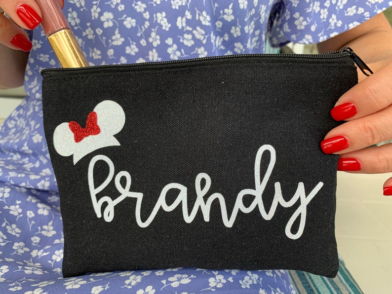 May include: Black canvas cosmetic bag featuring a white Minnie Mouse design and the name "Brandy" in cursive. The bag has a black zip and holds a tube of lip gloss.