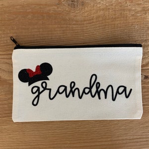 May include: A white canvas zippered pouch with the word "grandma" in black script. Above the word is a black mouse ear design with a red glitter bow. The zipper is black.