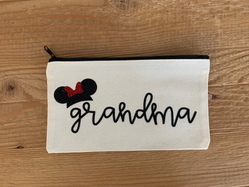 May include: A white canvas zippered pouch with the word "grandma" in black script. Above the word is a black mouse ear design with a red glitter bow. The zipper is black.