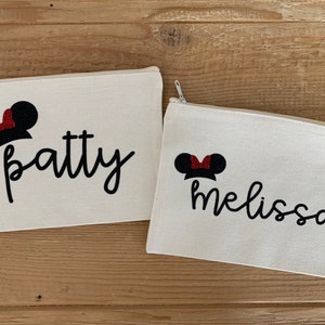 May include: Two off-white canvas zipper pouches. Each pouch features a black script name and a Minnie Mouse-inspired design with black ears and a red glitter bow. The names "Patty" and "Melissa" are displayed on the pouches.