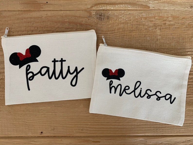 May include: Two off-white canvas zipper pouches. Each pouch features a black script name and a Minnie Mouse-inspired design with black ears and a red glitter bow. The names "Patty" and "Melissa" are displayed on the pouches.