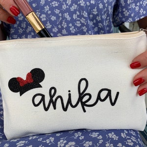 May include: A white canvas makeup bag featuring a black glitter Minnie Mouse design and a red glitter bow. The name "ahika" is written in black cursive script. A makeup brush is visible inside the bag.