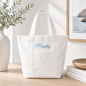 May include: A white canvas tote bag featuring two sturdy handles and a front exterior pocket. The name Murphy is embroidered in a light blue cursive script on the front of the bag, centered above the pocket. The bag has a structured base.