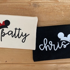 May include: Two canvas zipper pouches. One is off-white with "patty" in black script and a Minnie Mouse silhouette. The other is black with "chris" in white script and a Mickey Mouse silhouette. Both pouches are on a wooden surface.