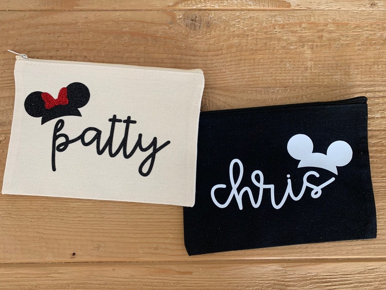 May include: Two canvas zipper pouches. One is off-white with "patty" in black script and a Minnie Mouse silhouette. The other is black with "chris" in white script and a Mickey Mouse silhouette. Both pouches are on a wooden surface.