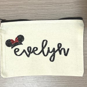 May include: A cream canvas pouch with a black zip, featuring the name "evelyn" in black script. A black glitter Mickey Mouse ear design with a red bow is on the upper left corner.