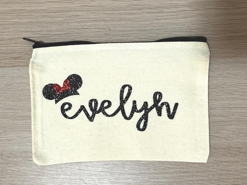 May include: A cream canvas pouch with a black zip, featuring the name "evelyn" in black script. A black glitter Mickey Mouse ear design with a red bow is on the upper left corner.