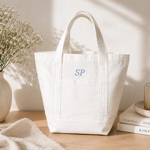 May include: A white canvas tote bag with a rectangular shape and two handles. The bag has a front pocket with the embroidered initials "SP" in light blue. A vase of white flowers, a candle, and books are also in the image.