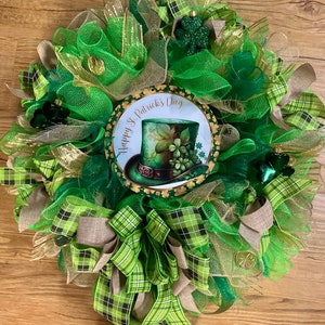 May include: A festive St. Patrick's Day wreath with green, gold, and tan mesh ribbons. The wreath features a central circular design with a leprechaun hat and the text "Happy St. Patrick's Day". Decorative shamrocks and gold coins are also included.