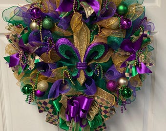 May include: A Mardi Gras wreath with gold, green, and purple mesh, ribbons, and ornaments. A gold, green, and purple fleur-de-lis is in the center.