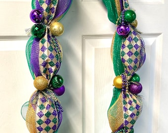 May include: A decorative Mardi Gras garland featuring green, gold, and purple accents. The garland is adorned with decorative balls in various sizes and colors, along with beaded accents. The design incorporates diamond patterns and ribbons.