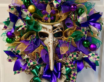 May include: A festive Mardi Gras wreath featuring a white mask with a long nose, surrounded by layers of purple, green, and gold mesh. Embellished with decorative bows, beads, and ornaments in matching colors. A vibrant and celebratory decoration.