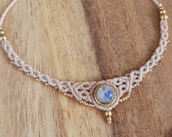 May include: A handmade macrame necklace with a central moonstone and gold beads. The beige cord necklace is displayed on a wooden tray with seashells and flower petals.