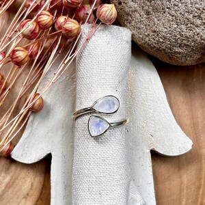 Könnte beinhalten: Ein silberner Ring mit zwei tropfenförmigen Mondsteinen. Der Ring wird auf einem weißen, handförmigen Halter mit einem Leinentuch präsentiert. Getrocknete Blumen und ein Stein befinden sich im Hintergrund, auf einer Holzoberfläche.