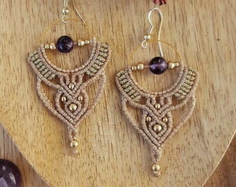 May include: A pair of handmade macrame earrings with gold-colored accents and a dark purple bead. The earrings are displayed on a wooden surface with decorative stones and dried seed pods.