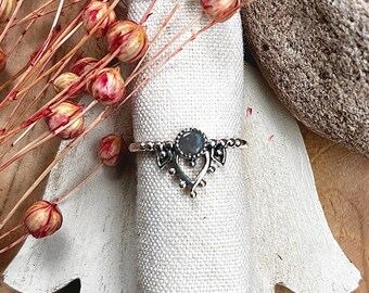 Op de afbeelding: Een zilveren ring met een ovale, grijze edelsteen wordt gepresenteerd op een witte stof. De ring heeft een sierlijk ontwerp met decoratieve elementen. De ring wordt gepresenteerd op een witte, handvormige houten standaard, met gedroogde bloemen en een steen op de achtergrond.