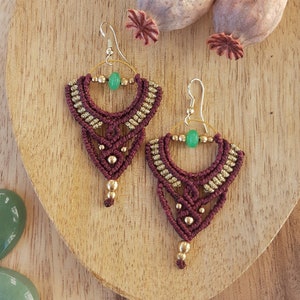 May include: A pair of handmade macrame earrings in a deep burgundy color, accented with gold beads and a green gemstone. The earrings are displayed on a wooden surface with decorative elements.