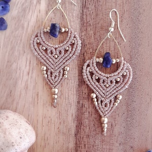 May include: A pair of handmade macrame earrings with a beige woven design, gold-colored accents, and a dark blue gemstone. The earrings are displayed on a wooden surface with scattered blue gemstones and dried flowers.