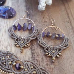 May include: A jewelry set featuring a macrame bracelet and hoop earrings. The earrings and bracelet are adorned with purple gemstones and gold-colored beads. Several polished purple stones are also visible.