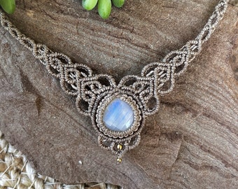 May include: A handcrafted macrame necklace featuring a central, faceted, light blue moonstone. The necklace is made with tan-colored cord, with intricate knotting patterns. Small gold-colored beads accent the design. The necklace is displayed on a brown, textured surface.