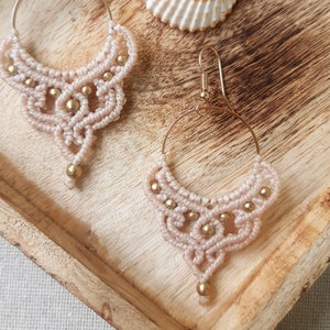 May include: A pair of handmade macrame earrings with a light pink woven design and gold-colored accents. The earrings are displayed on a wooden tray next to a seashell. The earrings have a circular hoop design.