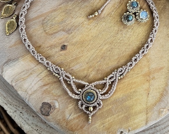 May include: A handcrafted macrame necklace with a central blue gemstone pendant and matching stud earrings. The necklace and earrings are made with beige cord and accented with gold-colored beads. The jewelry is displayed on a wooden surface.