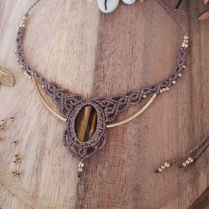 May include: A handcrafted macrame necklace with a large oval tiger's eye stone pendant. The necklace features a brown cord with gold-colored beads and a gold-toned metal accent. The design is bohemian and natural, with a focus on earthy tones and textures.