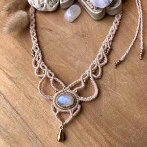 May include: A beige macrame necklace with a central oval moonstone and a gold-colored teardrop pendant. The necklace features intricate knotting and small gold beads. The background includes decorative coral and a wooden surface.