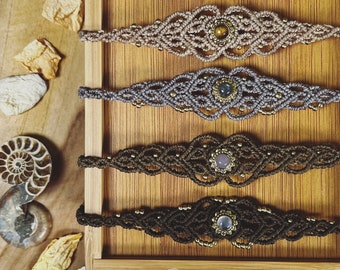 May include: Six macrame bracelets in varying colors, each featuring a central gemstone and gold-colored beads. The bracelets are displayed on a wooden tray, showcasing the intricate knotting patterns and earthy tones of the jewelry.