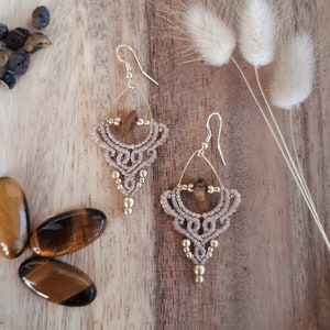 May include: A pair of handmade macrame earrings with gold-colored accents and a tiger's eye stone. The earrings feature a beige, woven design with small gold beads. The image also shows polished tiger's eye stones and small, dark blue stones.