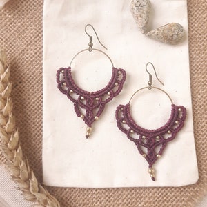 May include: A pair of handmade macrame earrings in a deep burgundy color. The earrings feature a circular brass hoop with intricate knotting and small gold beads. They are displayed on a white pouch against a neutral background.