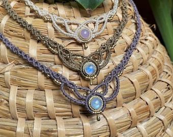 May include: Four macrame necklaces in shades of cream, olive green, and purple, each featuring a central gemstone pendant. The necklaces are adorned with small gold beads and are displayed on a woven basket.