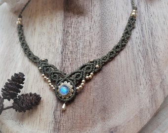 May include: A handcrafted macrame necklace in olive green with gold-colored beads. The focal point is a round, iridescent blue gemstone pendant with a gold-colored teardrop charm. The necklace is displayed on a wooden surface.