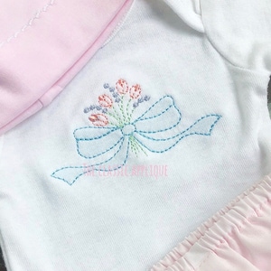 May include: White baby onesie with a blue embroidered bow and a bouquet of flowers in pink, orange, and purple.