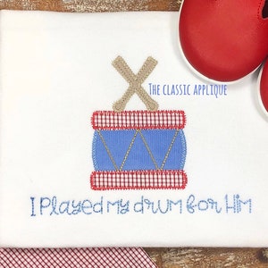 May include: White onesie with a blue and red drum applique and the text "I Played My Drum For Him". The drum has a gold and blue design and a brown stick.