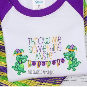 Könnte beinhalten: Ein weißes und violettes T-Shirt in Kindergröße mit einem grünen Alligator, der eine Perlenkette hält. Der Text "Throw Me Something Mister" ist auf das Shirt gestickt. Der Text "The Classic Applique" ist ebenfalls auf das Shirt gestickt.