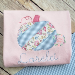 May include: A pink long-sleeved shirt with a patchwork pumpkin applique and the name "Lorelei" embroidered in blue thread.