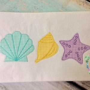 May include: A white t-shirt with three embroidered appliques: a teal seashell, a yellow seashell, and a purple starfish.