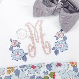 Peut inclure: Un body blanc avec un motif brodé de fleurs roses et la lettre "M" en écriture cursive. Le body est associé à un nœud gris et un tissu à motifs floraux.