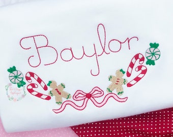 Op de afbeelding: Wit shirt met lange mouwen met de rode geborduurde naam "Baylor" en een kerstsnoep- en peperkoekmannetjesontwerp. Het shirt heeft een gerimpelde halslijn en is geplaatst tegen een rood-roze stippenachtergrond.