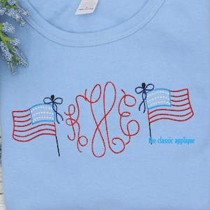 May include: Light blue t-shirt with a red, white, and blue embroidered design featuring two American flags and a monogram. The text "the classic applique" is also embroidered on the shirt.