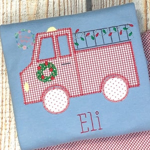 May include: A red and white gingham truck with a wreath on the side and Christmas lights on the back. The truck is embroidered on a light blue shirt with the name "Eli" embroidered below it.