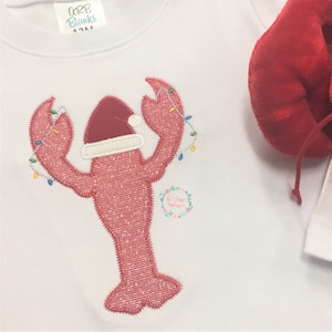 May include: A white baby onesie with a red embroidered lobster wearing a Santa hat and holding Christmas lights. The onesie has a small tag that says "CCB Blanks 12M".