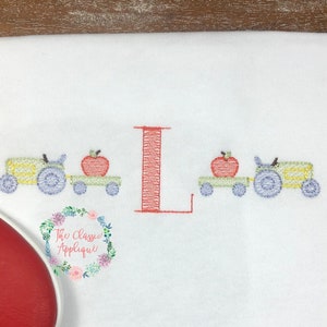 Op de afbeelding: Geborduurde afbeelding van een tractor die een wagen met een appel trekt op een witte stof. De letter L is in rood geborduurd in het midden van het ontwerp.