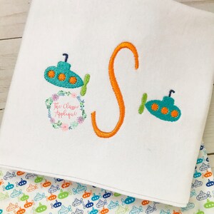 May include: A white fabric with an orange letter S and two blue and green submarine appliques. The fabric has a small floral wreath with the text "The Classic Applique". The fabric is folded over a white fabric with a repeating pattern of blue, green, and orange submarines.