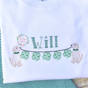 May include: White t-shirt with a green shamrock garland and two golden retriever dogs on either end. The name "Will" is embroidered in the center of the garland.