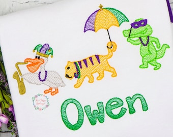 May include: White t-shirt with a green, yellow, and purple embroidered design featuring a pelican playing a saxophone, a tiger, an alligator wearing a mask, and an umbrella. The name "Owen" is embroidered in green below the design.