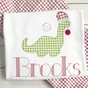 May include: A white onesie with a green and white gingham dinosaur wearing a red and white gingham Santa hat and holding a red ornament. The onesie has the name "Brooks" embroidered in red on the bottom.